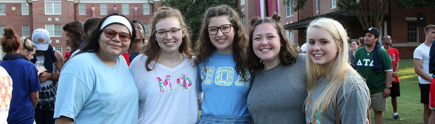 Five students shoulder to shoulder facing the camera in a crowd of other students